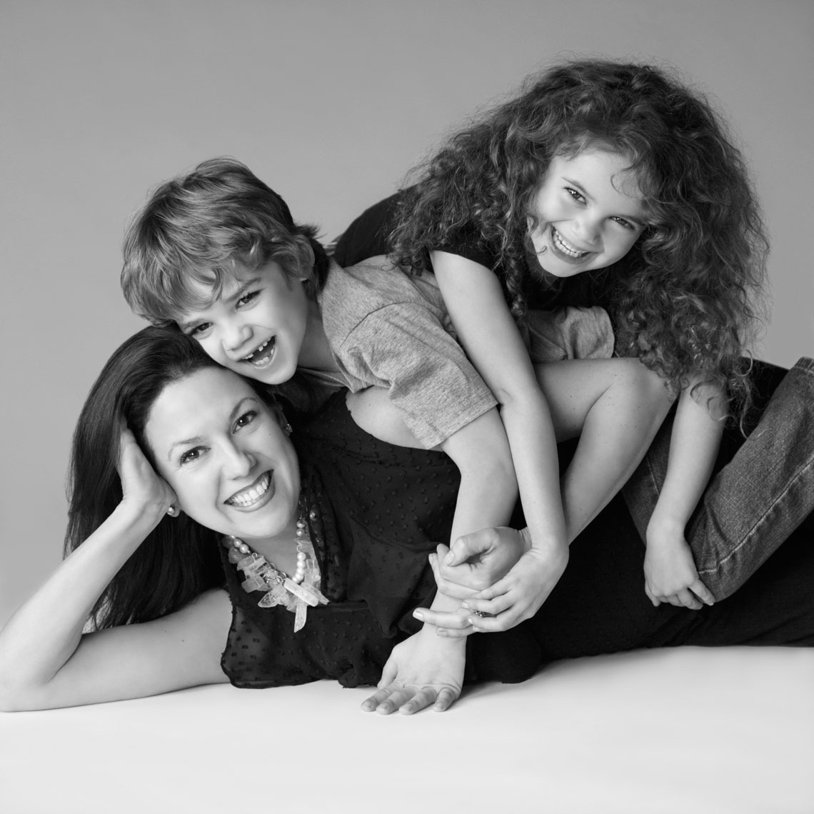 Black and white family portrait of Noel Ledoux and her children lying on each other on the background in the studio in Dallas,  Texas, by photographer John Derryberry Photography
