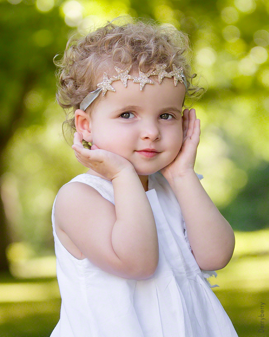 Portrait of a cute little curly headed blonde girl outside at a park in Highland Park, Texas with her hands on her face and wearing a headband with stars on it.