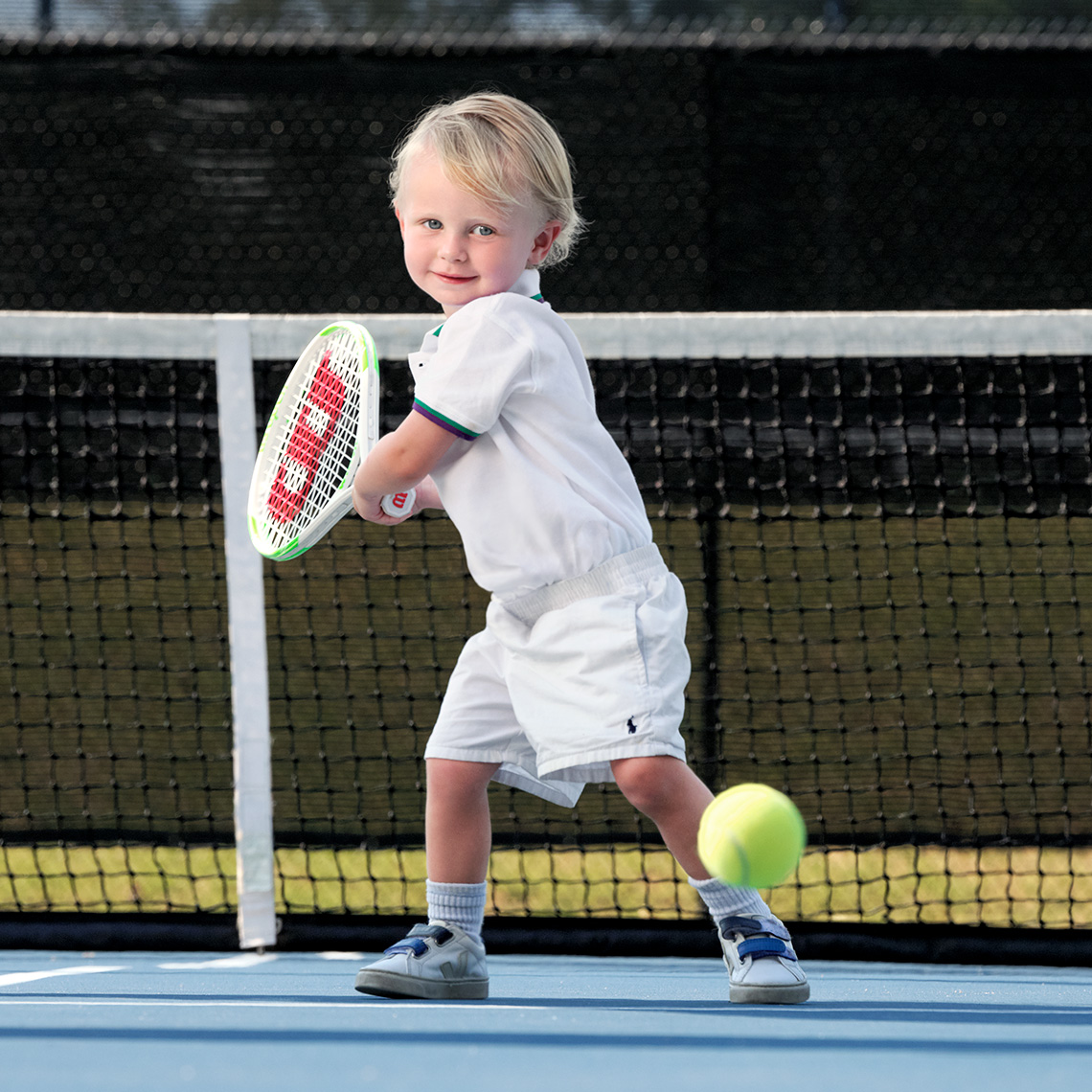Color portrait of a blonde  little boy in white swinging a tennis racket to hit a ball