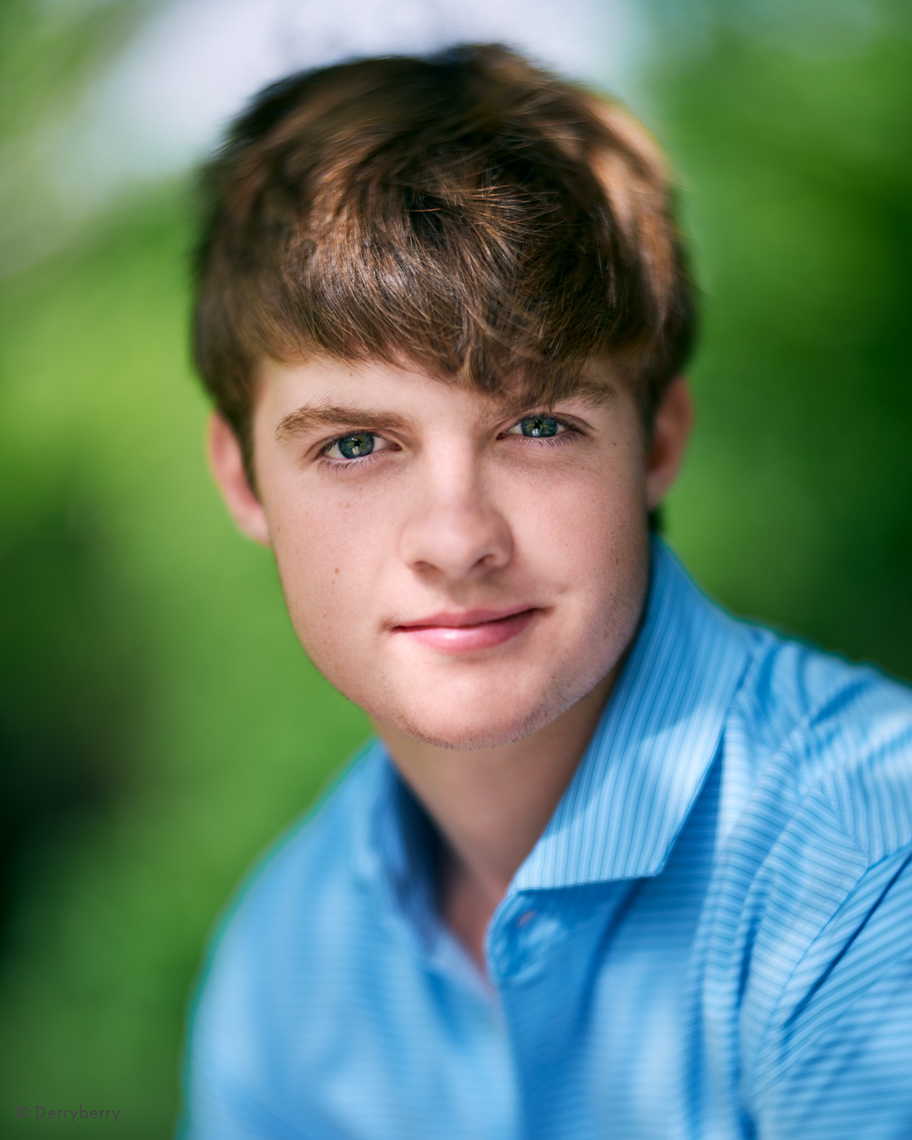 Headshot of senior boy in blue shirt outdoors at a park with shallow depth of field