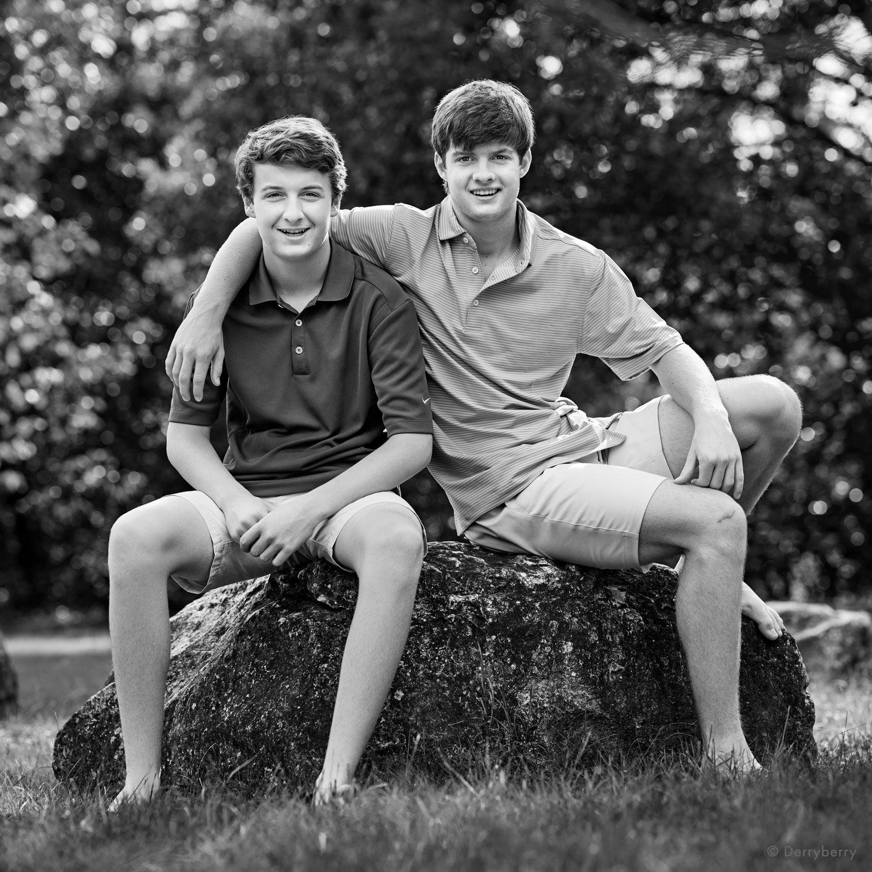 Two brothers, one a senior,  smiling and seated on a large rock with trees in the background