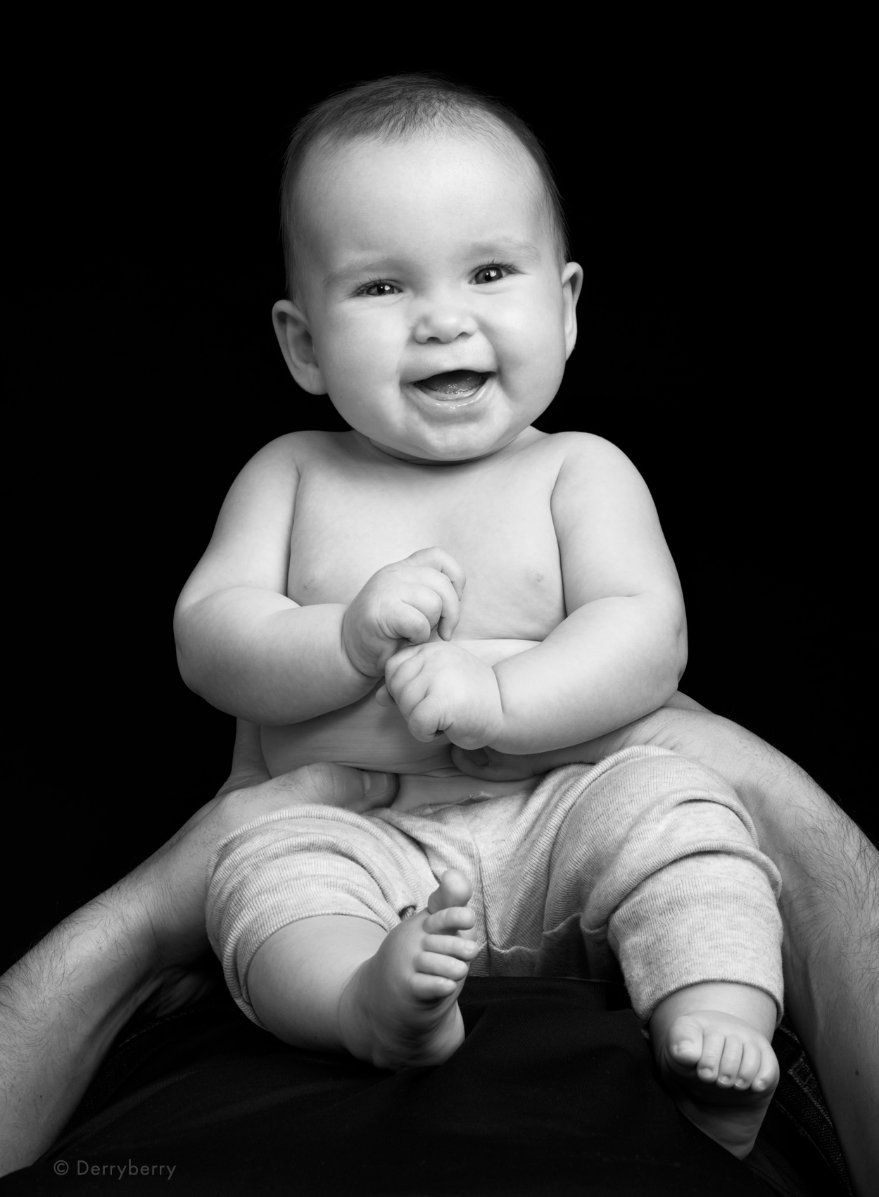 B&W portrait of a happy, smiling baby boy sitting on his dads tummy and with a black background
