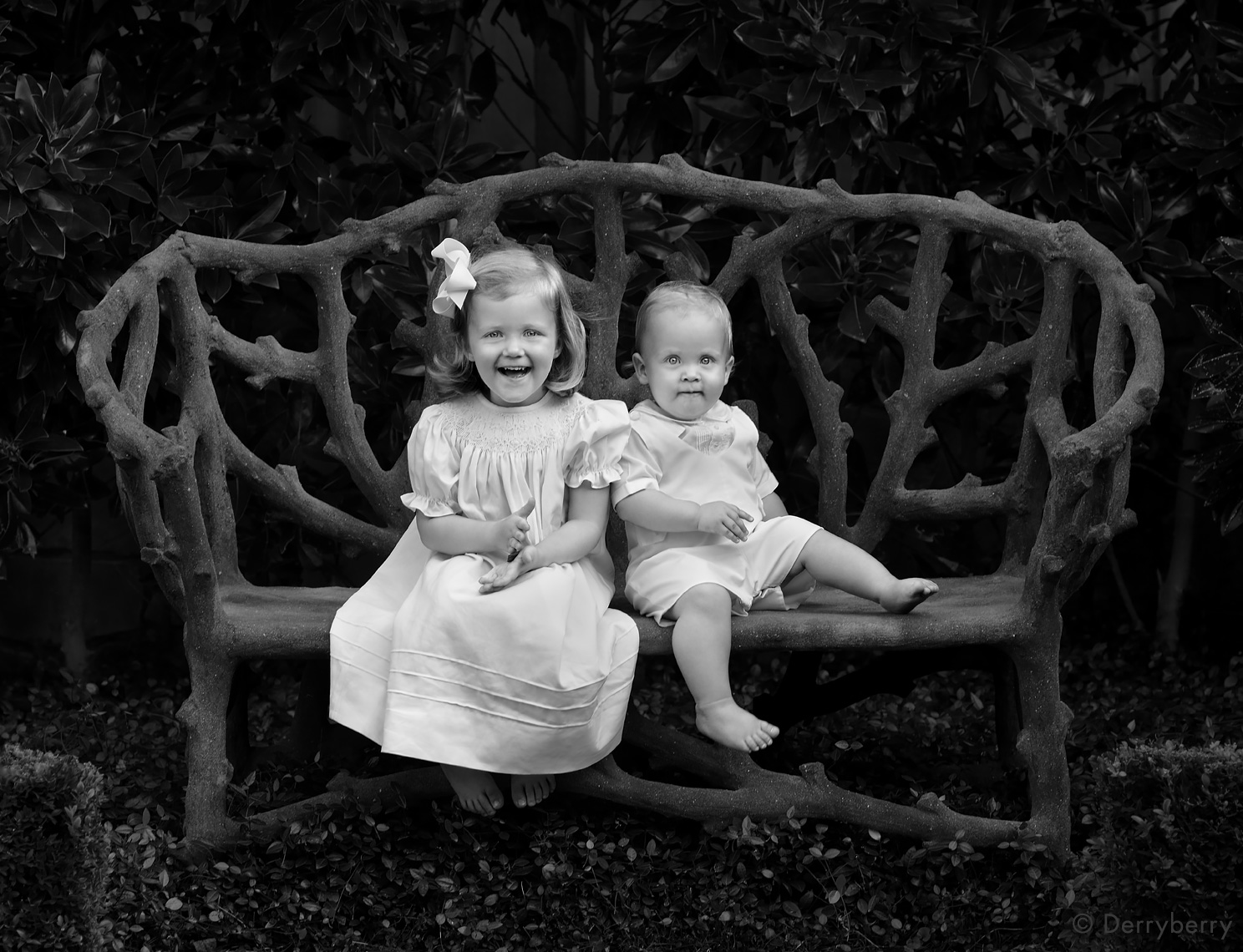 Black and white portrait of a young brother and sister sitting on a faux bois bench on location in a garden